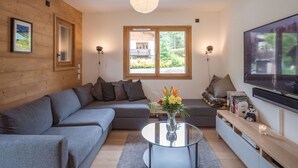 A modern living room with a sectional sofa, a glass coffee table, a flat-screen TV, and a large window with a view of greenery.