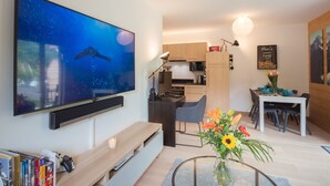 A modern living room with a flat-screen TV, a dining table with chairs, a kitchen area, and a bookshelf with books.
