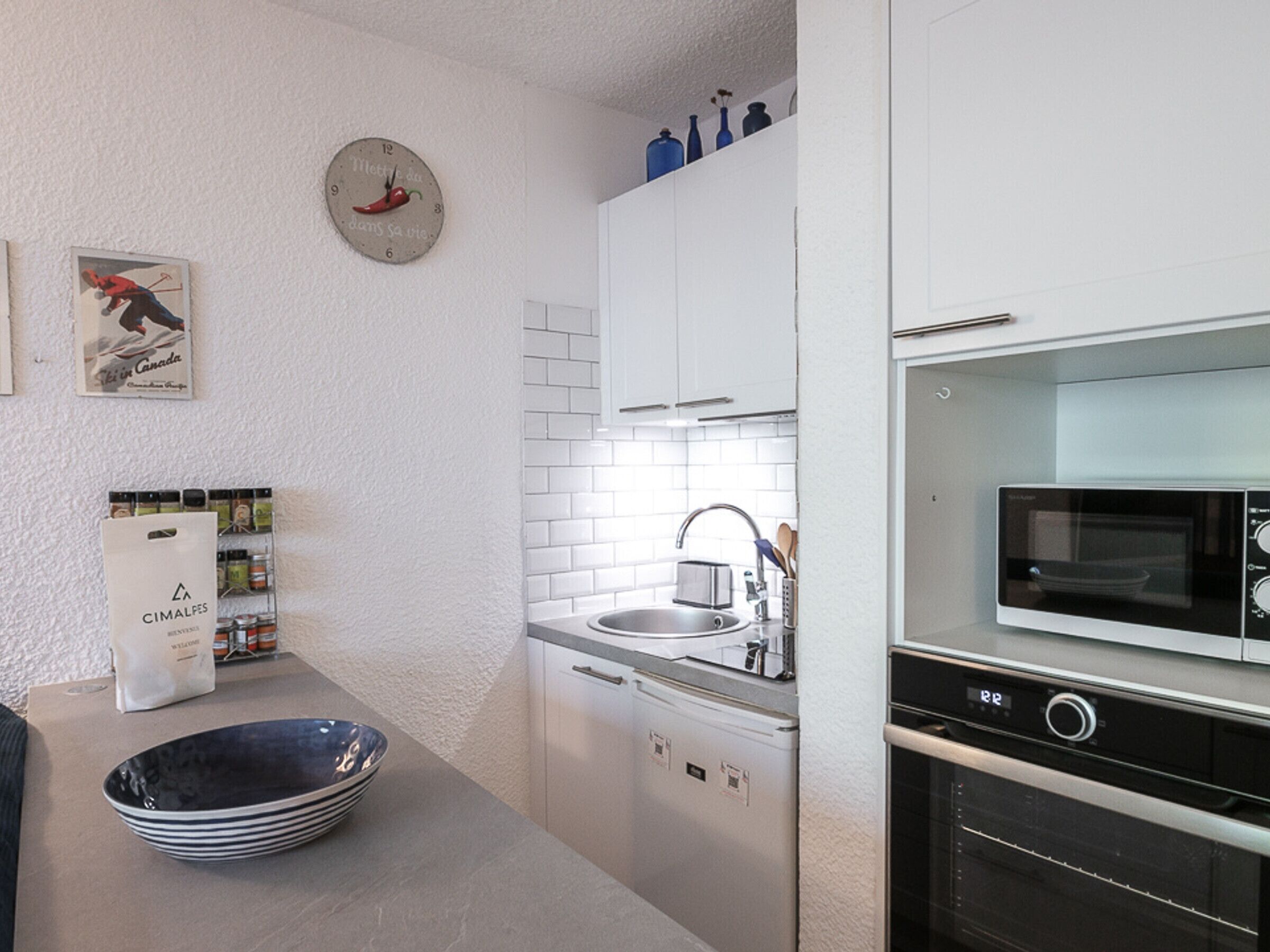 A modern kitchen with white cabinets, a microwave, an oven, a sink, and a clock with a chili pepper design.