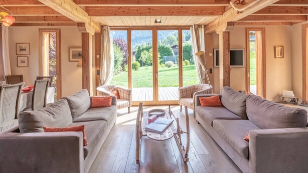 A cozy wooden interior with a sofa set, a dining table, and a view of a green landscape through large glass doors.