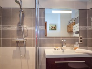 Ein modernes Badezimmer mit einer gläsernen Duschkabine, einem weißen Waschbecken mit Seifenschale und einem Spiegel, der ein Bild und einen Holzkorpus reflektiert.