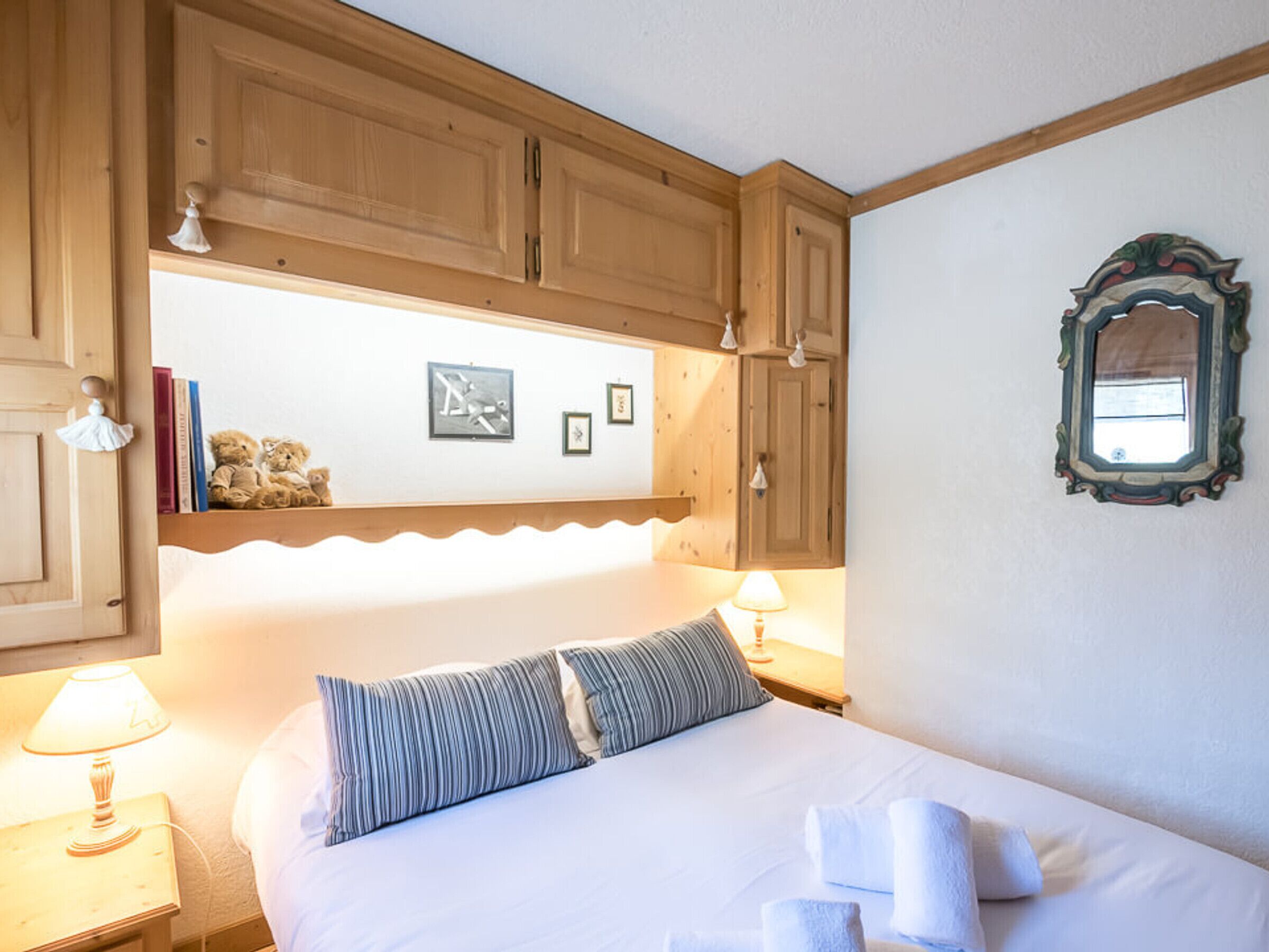 A bedroom with wooden cabinets, a bed with striped pillows, a bedside lamp, and framed pictures on the wall.