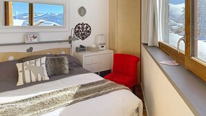 Quarto com cama, cadeira vermelha, guarda-roupa de madeira, secretária com abajur e janela com vista para neve.
