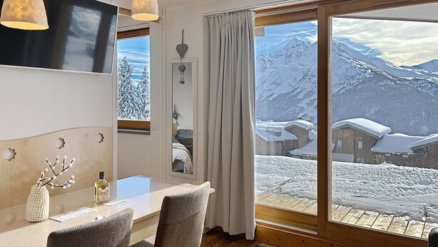 Um quarto com uma mesa de jantar de madeira, duas cadeiras, uma TV fixada na parede e uma janela grande com vista para uma montanha nevada.