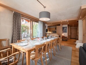 A dining area with a wooden table set for six, surrounded by chairs, with a view of a garden through large windows.