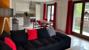 Un salon moderne avec un canapé noir, des coussins rouges et un coin repas comprenant une table en bois et des chaises rouges. On aperçoit à l’arrière-plan une cuisine avec des meubles blancs et une cuisinière à gaz.