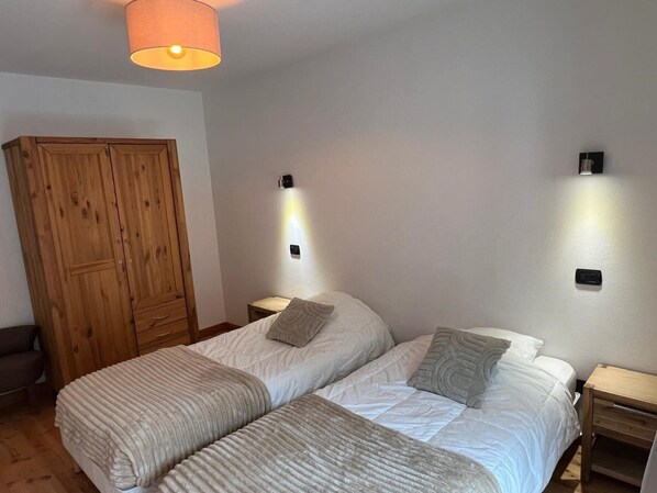 A bedroom with two beds, wooden wardrobe, nightstands, and wall-mounted lights.