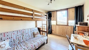 A cozy room with a wooden bunk bed, a sofa with patterned cushions, a dining table with chairs, and a window with a view of the outdoors.