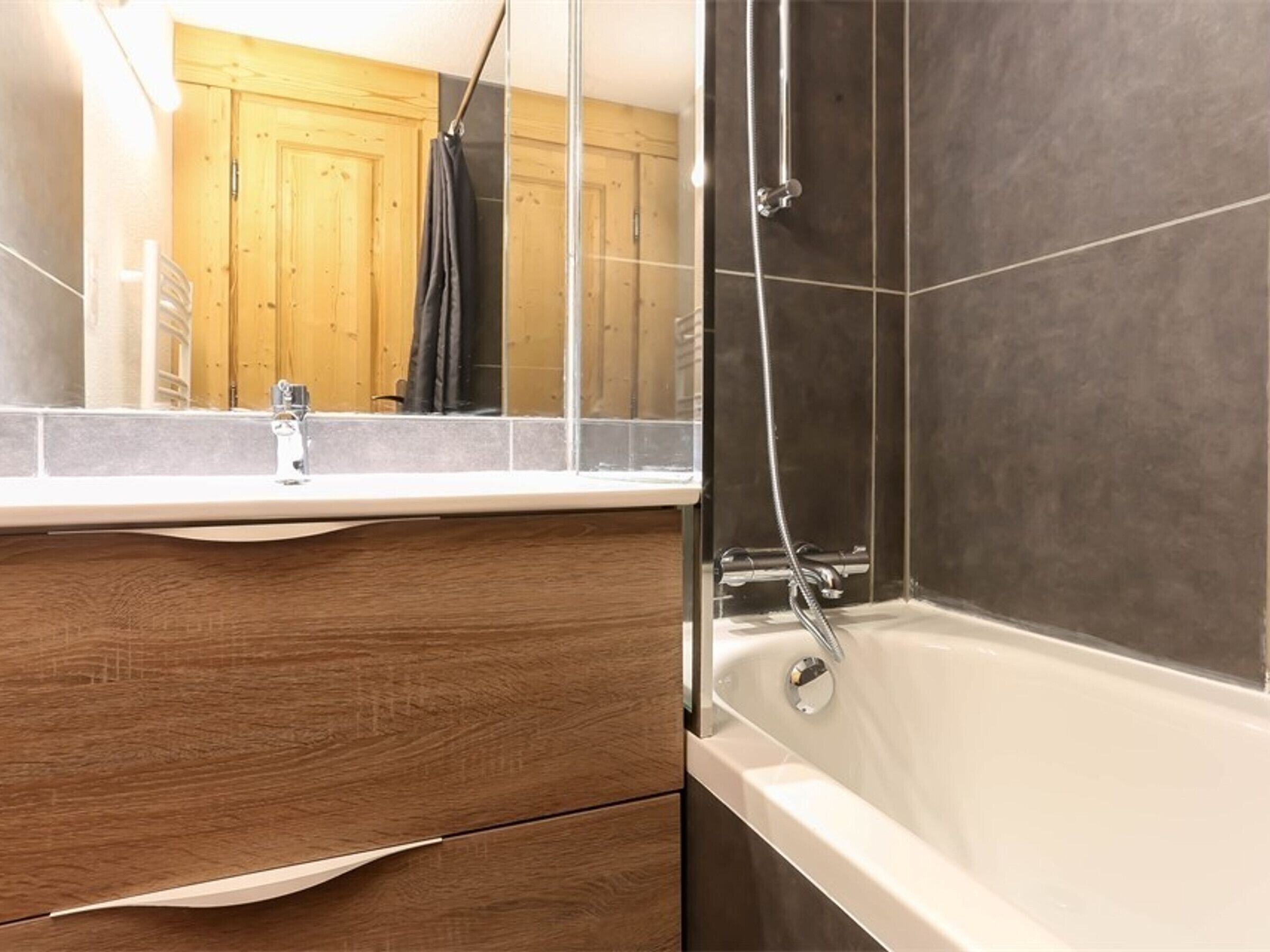 A bathroom with a wooden vanity, a white bathtub, and a mirror reflecting a wooden door and a black towel.