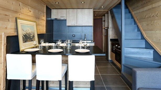 A dining area with a wooden ceiling, a kitchen in the background, and a staircase. - Renovated Duplex - Sleeps 6 - Ski-in/Ski-out - Mountain View Balcony - Close to Shops (Val Thorens)