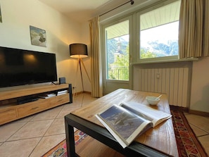 Une pièce avec une télévision, une lampe, une table sur laquelle se trouvent un livre et un bol, et une fenêtre donnant sur les montagnes.