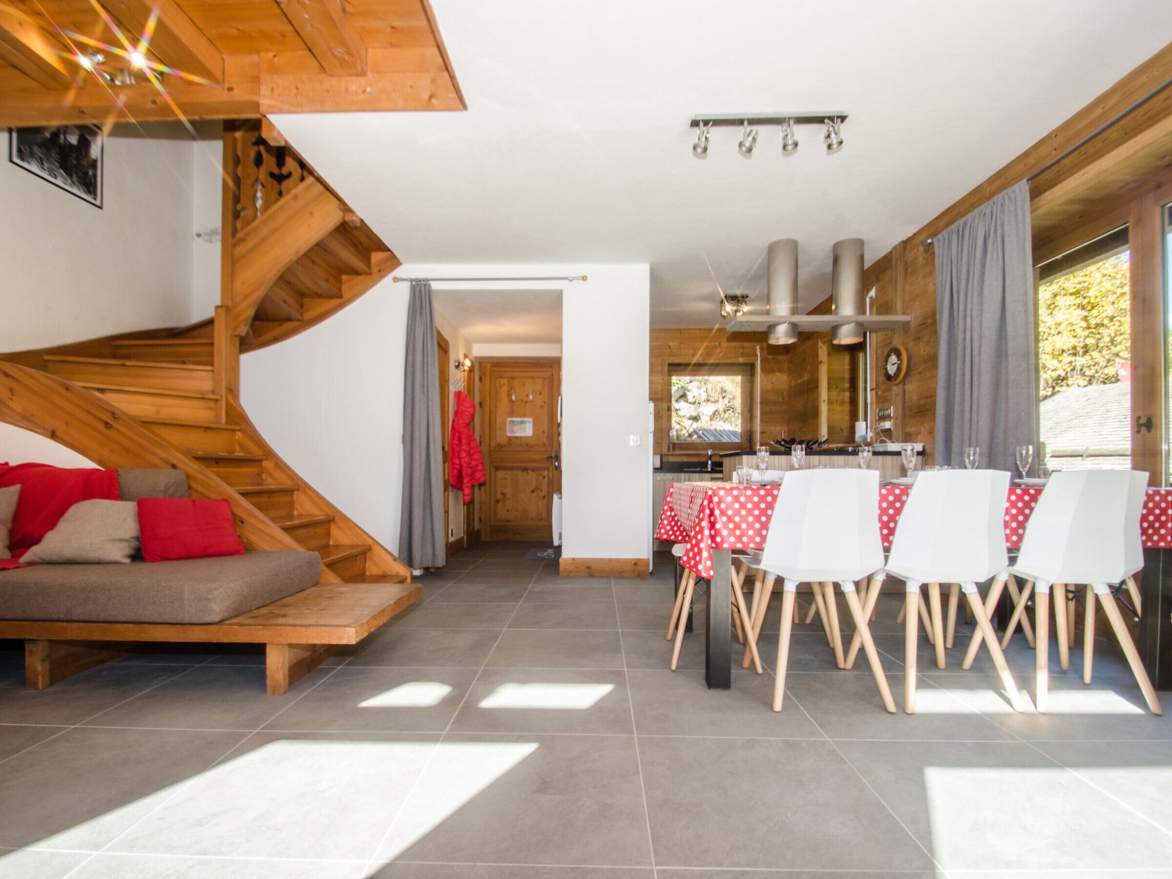 A modern interior with a wooden staircase, a dining area with a red and white checkered tablecloth, and a kitchen visible through an open door.
