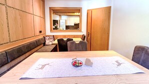 Ein gemütliches Zimmer mit einer hölzernen Sitzbank, einem Esstisch mit Unterlage und einer durch ein Fenster einsehbaren Küche.