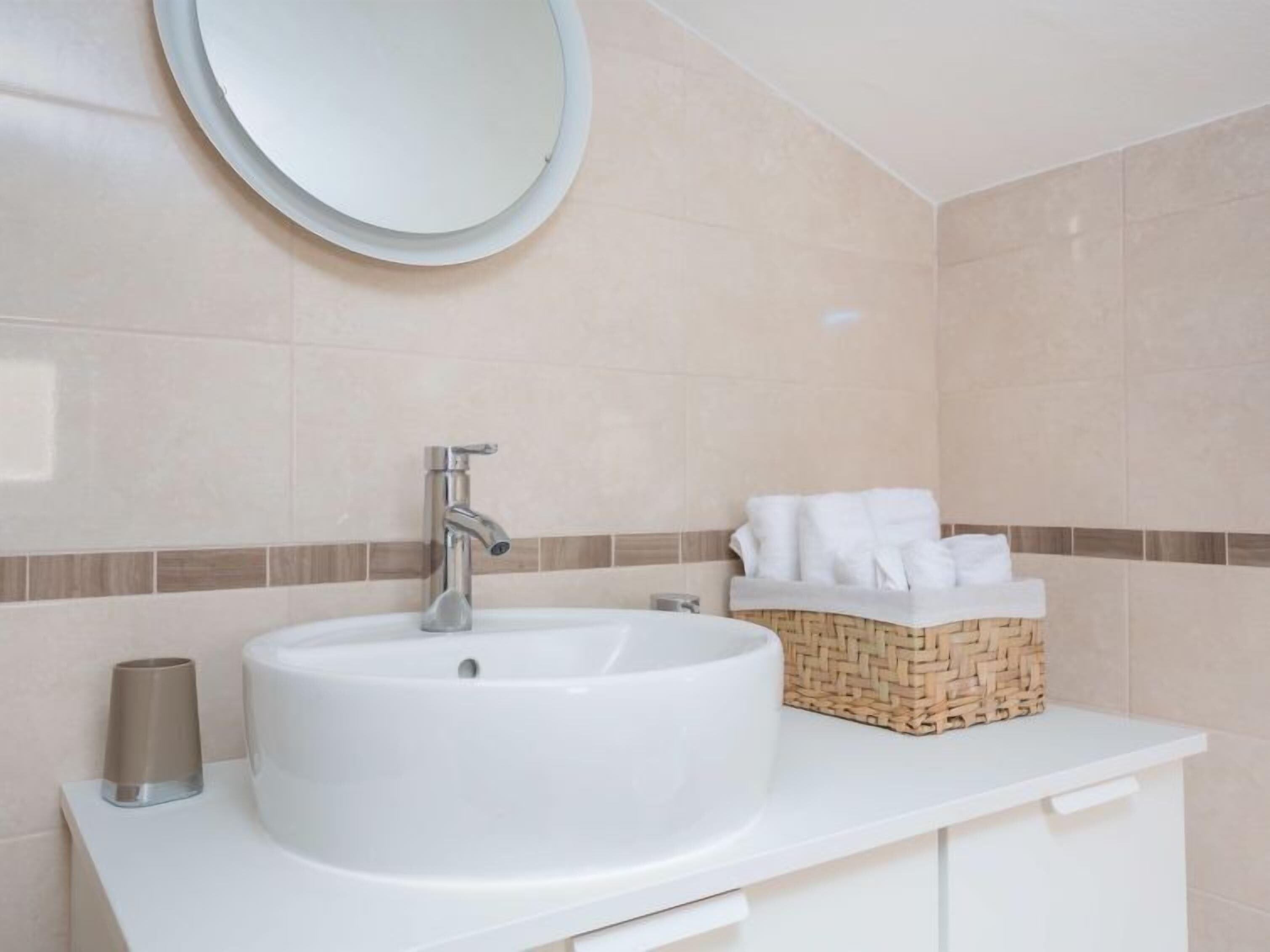 A bathroom with a round mirror, a white sink, a towel basket, and a toilet paper holder.