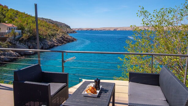 A balcony with wicker furniture, a tray of croissants, and a view of the sea and rocky coastline. - Wind Soleil by Interhome (Jablanac)