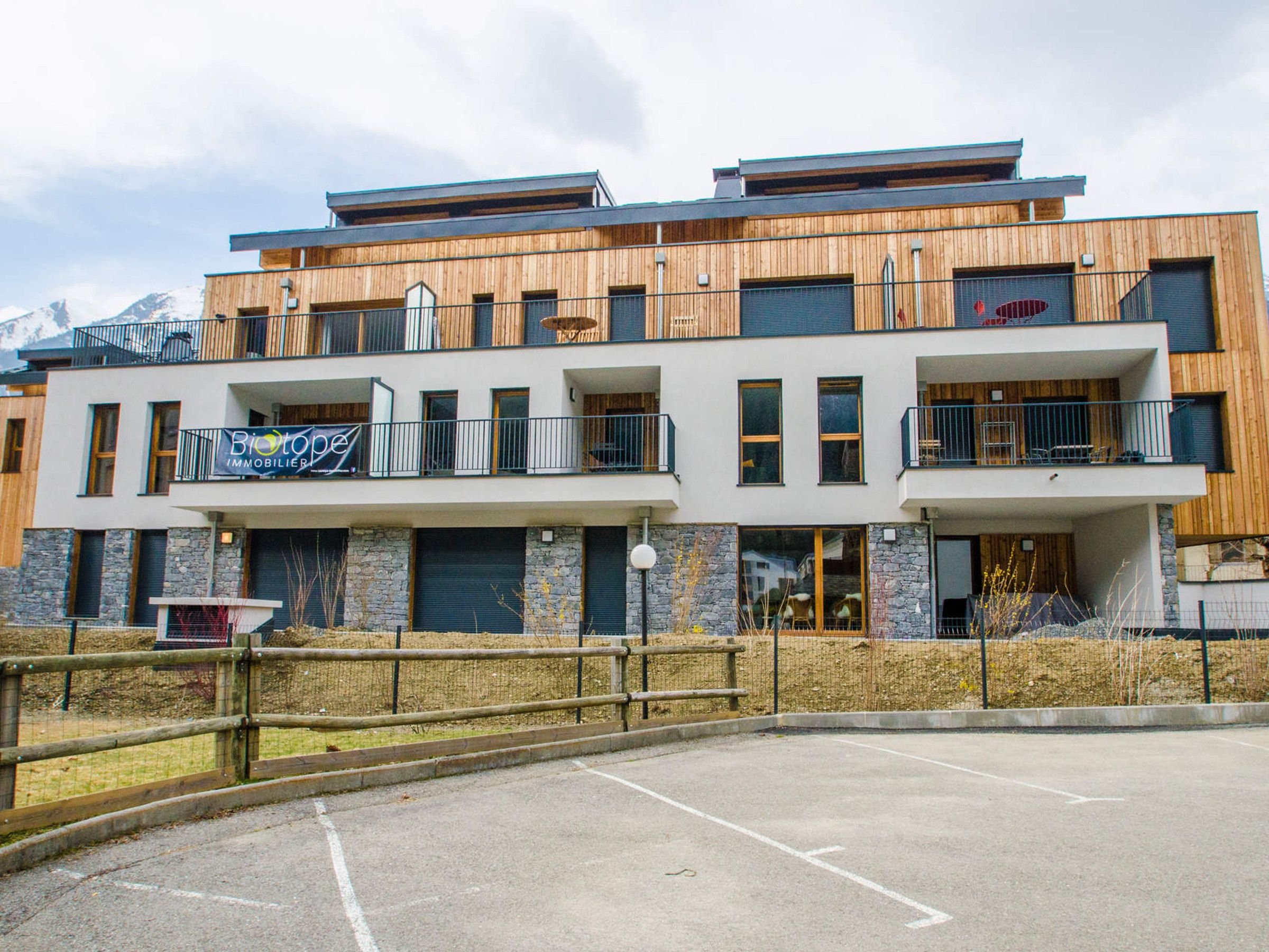 Un edificio moderno de varios pisos con fachada de madera, balcones y un cartel que dice 'Biotop Immobilien'.