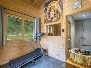 A wooden cabin interior with a treadmill, a large painting, a shower, and a view of greenery through the windows.