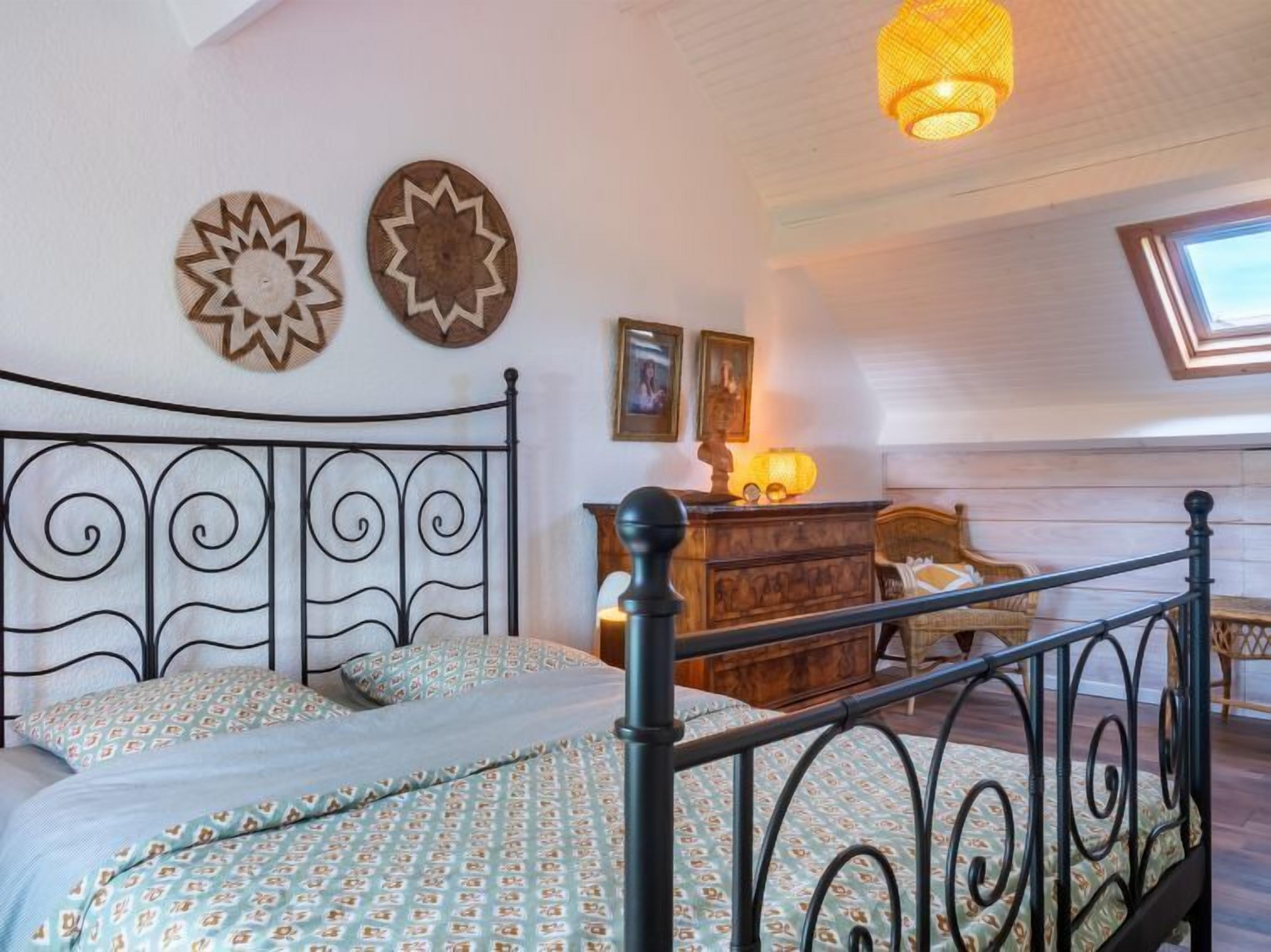 A bedroom with a metal bed frame, a patterned bedspread, a wooden dresser, a lamp, and two wall decorations.