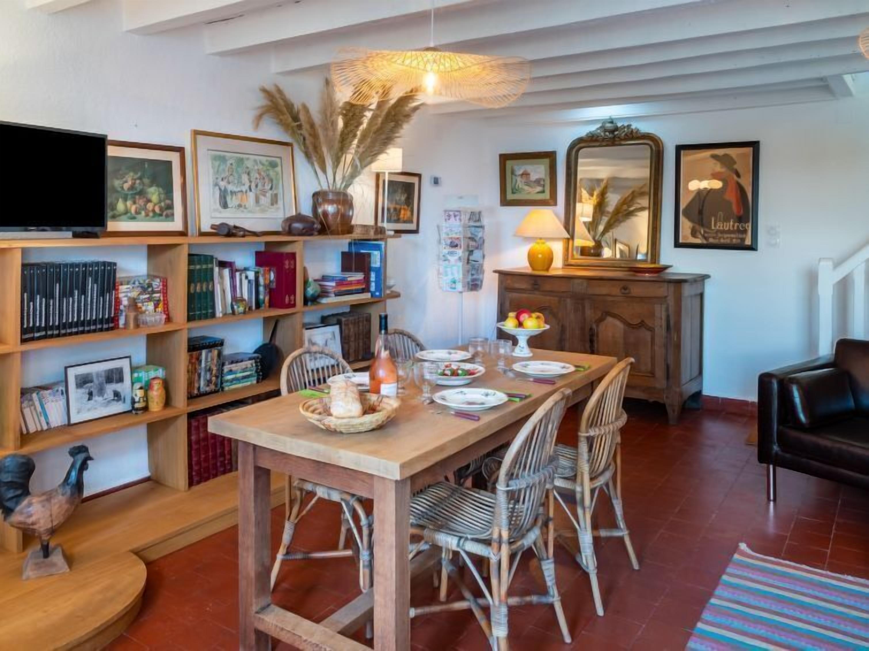 Een gezellig eetgedeelte met een houten tafel voor vier personen, omringd door rieten stoelen. Een boekenplank gevuld met boeken en decoratieve objecten bevindt zich aan de linkerkant, en boven de tafel hangt een spiegel.