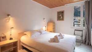 A bedroom with a wooden ceiling, a bed with white linens, a bedside lamp, a nightstand, a window with a view of buildings, and a painting on the wall. - Bright apartment for 5, Chamonix center, parking included (Chamonix-Mont-Blanc)