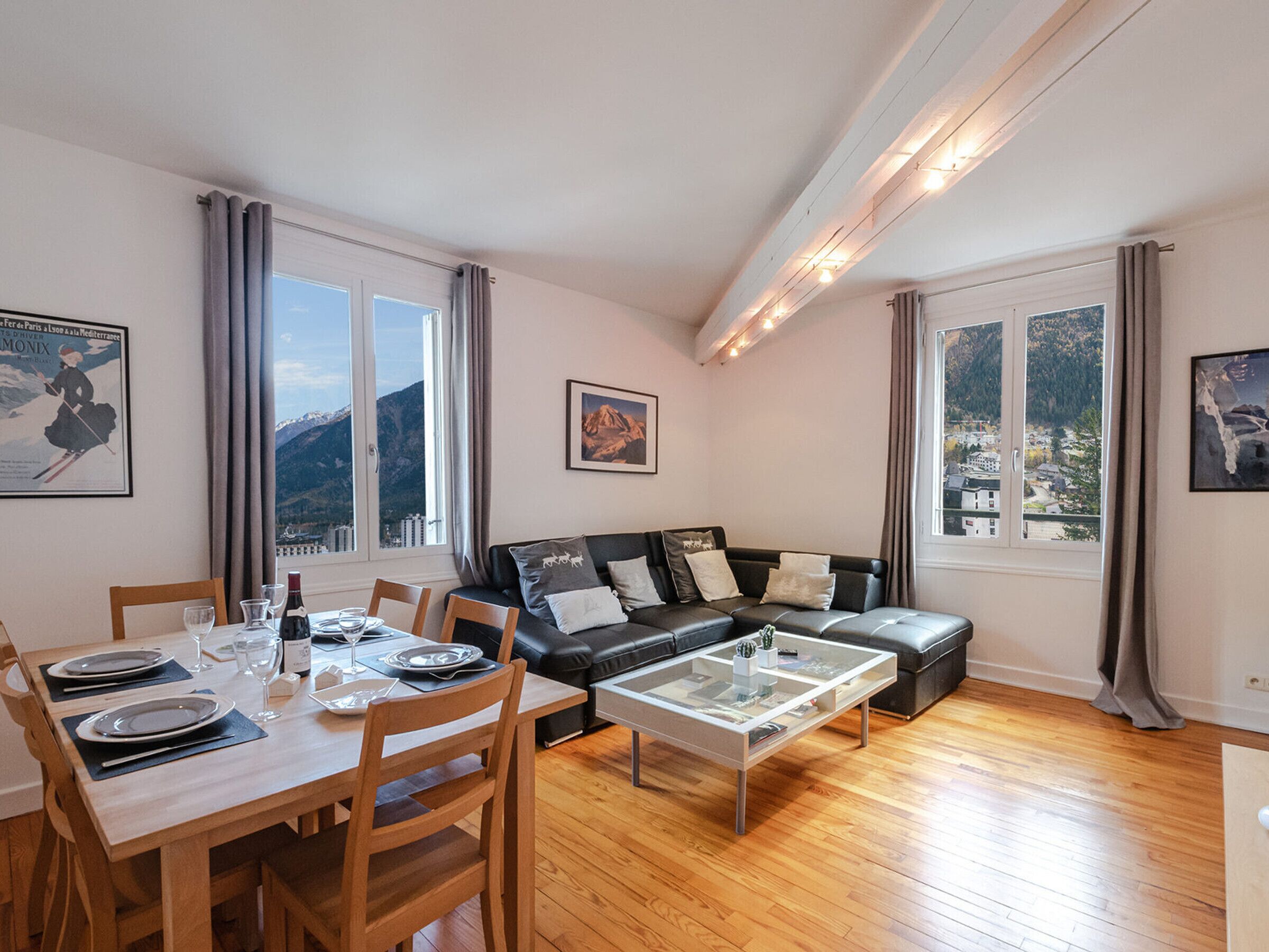 Een moderne woonkamer met een houten eettafel gedekt voor twee personen, een zwarte leren bank en een glazen salontafel. Grote ramen bieden uitzicht op een bergachtig landschap.