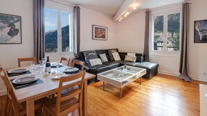 A modern living room with a wooden dining table set for two, a black leather sofa, and a glass coffee table. Large windows offer views of a mountainous landscape. - Bright apartment for 5, Chamonix center, parking included (Chamonix-Mont-Blanc)