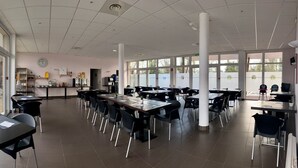 Un vaste espace intérieur aménagé pour les repas, avec de nombreuses tables et chaises, de grandes fenêtres et un coin cuisine à l’arrière-plan.