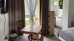 Une pièce avec une table et des chaises en bois, un téléviseur fixé au mur, un balcon avec vue et, au fond, une petite cuisine.