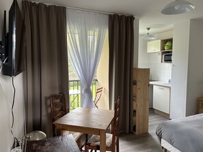Ein Zimmer mit Holztisch und -stühlen, einem an der Wand montierten Fernseher, einem Balkon mit Blick und einer kleinen Küchenzeile im Hintergrund.