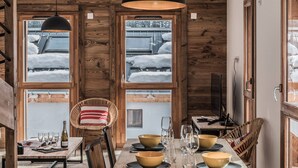 A dining area with a wooden table set for four, surrounded by chairs. There are two pendant lights above the table, and a window with a view of snow-covered roofs outside. - 3-star duplex for 6 pers, Chamonix, private parking (Chamonix-Mont-Blanc)