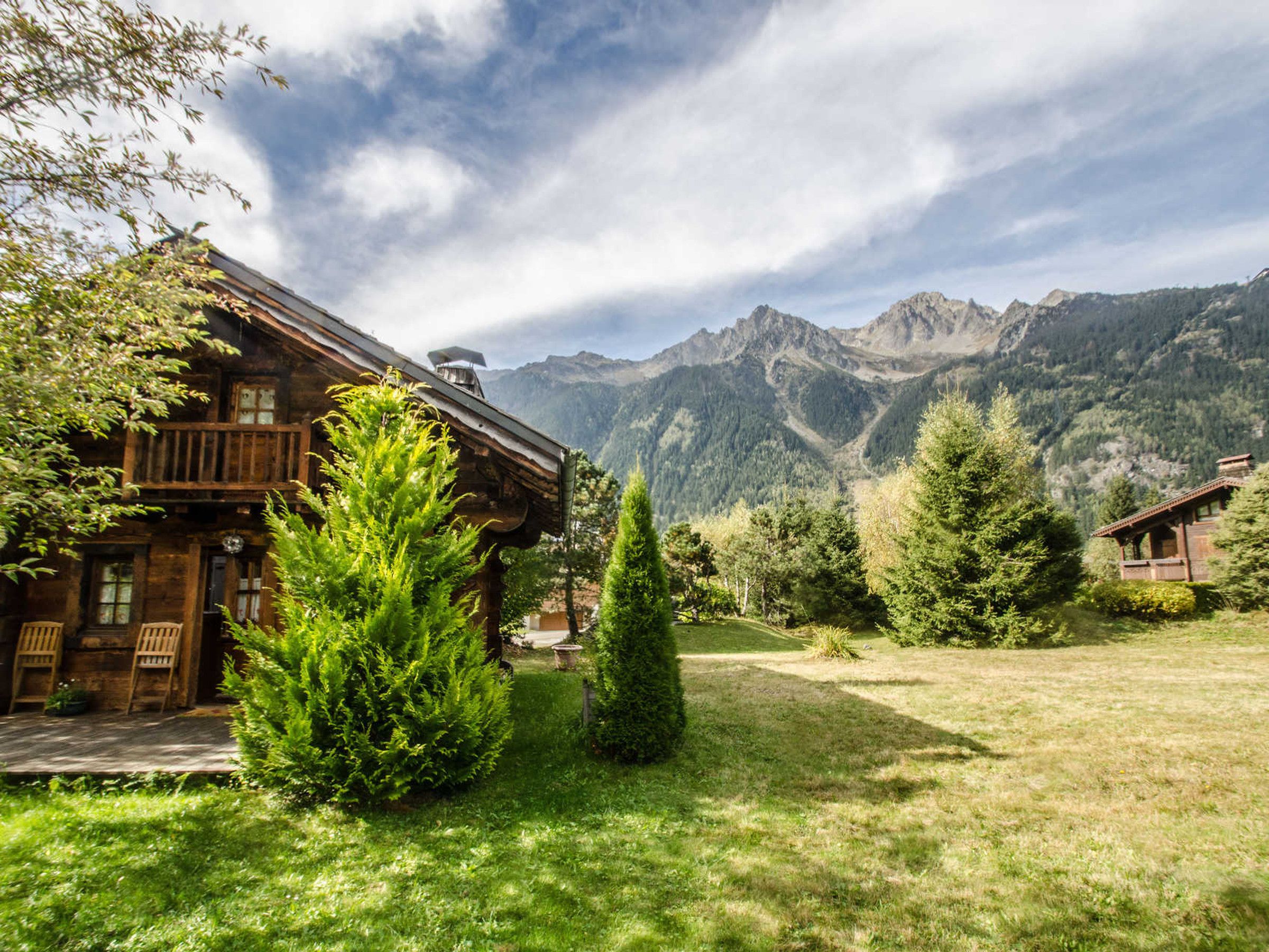 A wooden cabin with a balcony, surrounded by lush greenery and mountains in the background.
