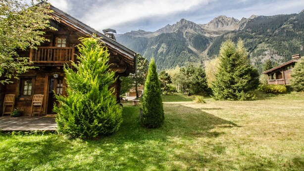 Una cabana de fusta amb un balcó, envoltada de vegetació abundant i muntanyes al fons.