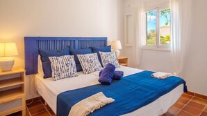 A neatly made bed with blue bedding and striped pillows, a bedside lamp, and a window with curtains. - Murada Lisboa by Interhome (Cala Murada)