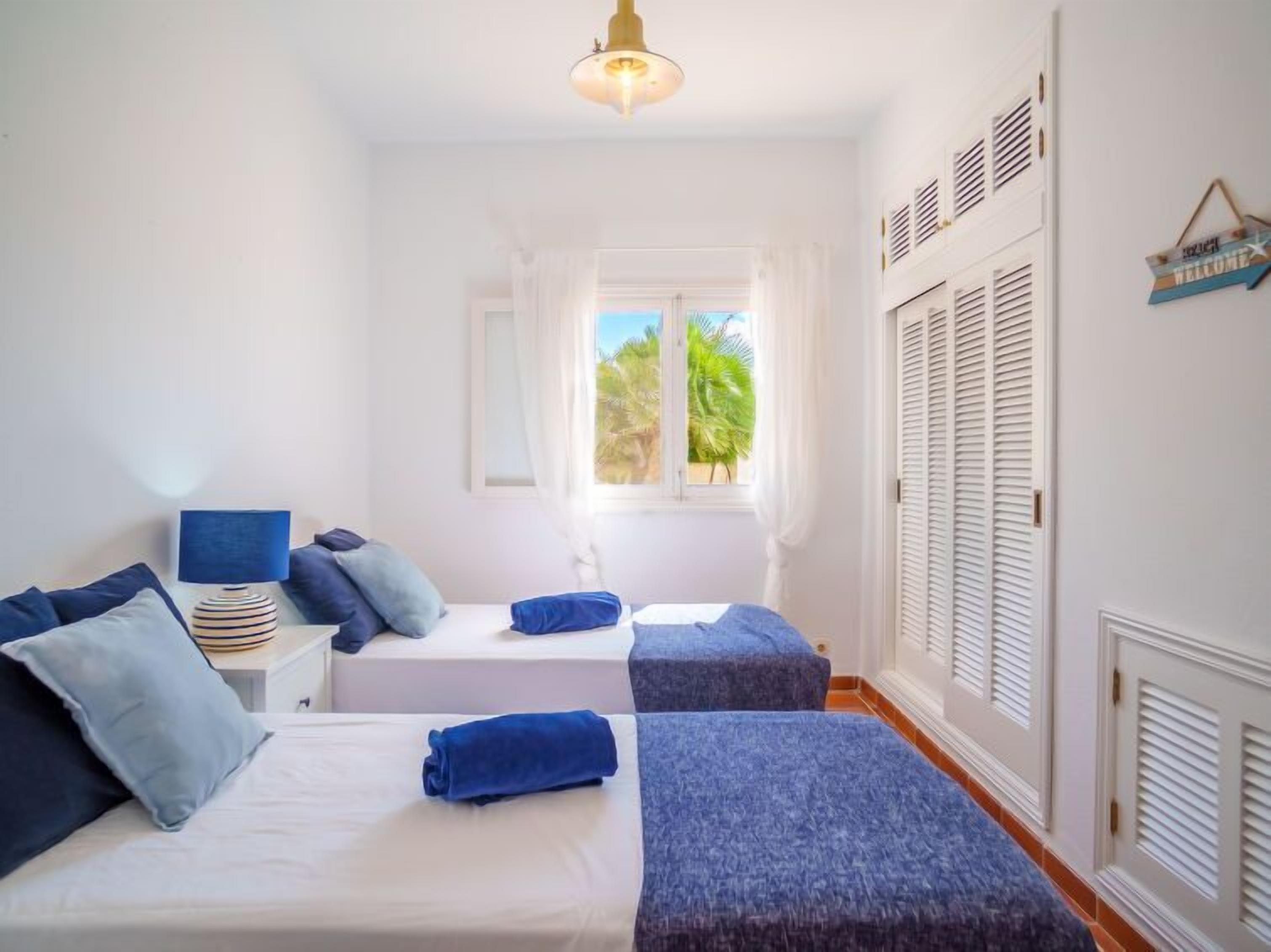 A bedroom with two beds, a bedside table, a lamp, a window with curtains, and a view of greenery outside.