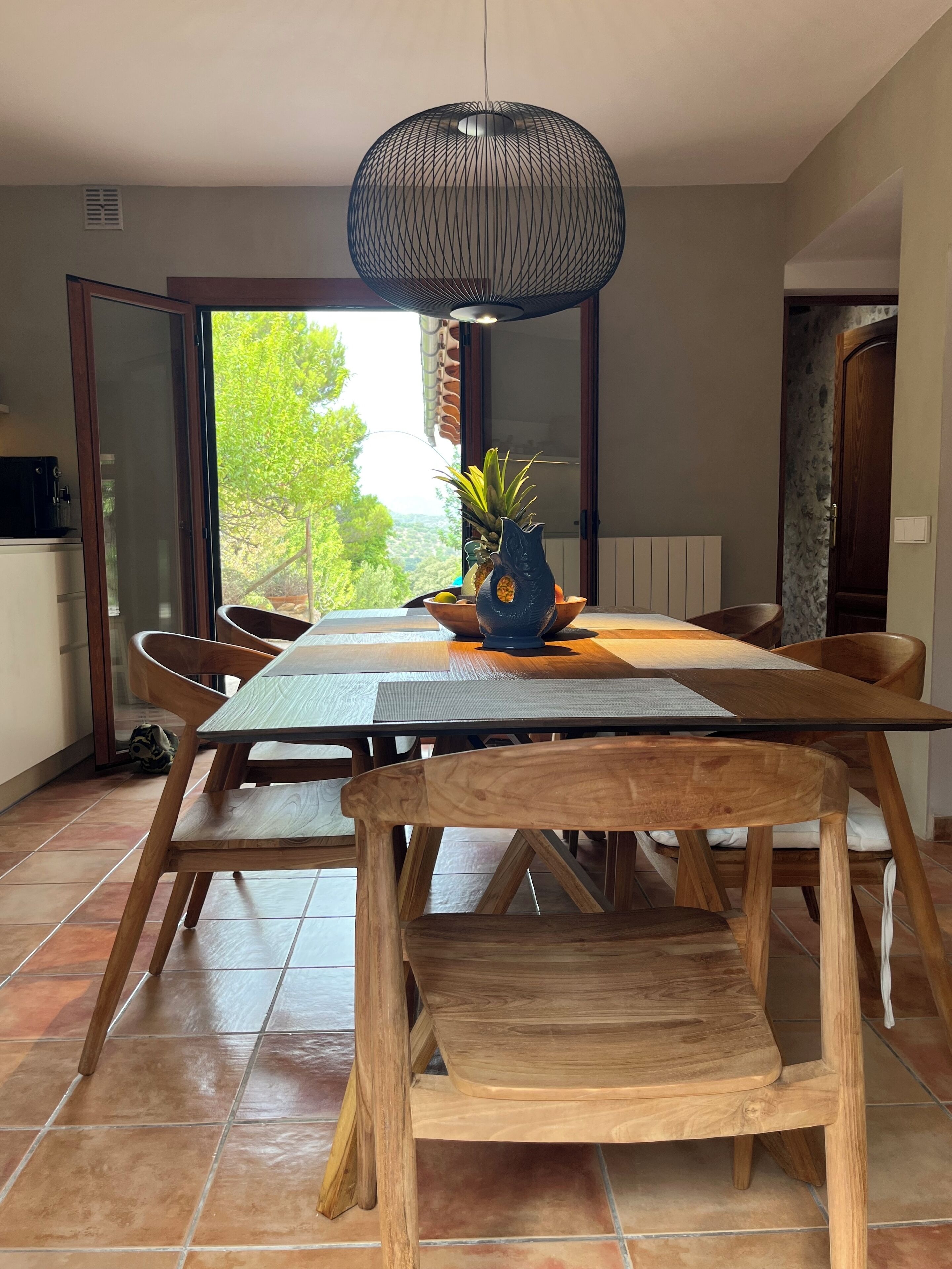 Un espace repas avec une table et des chaises en bois, un luminaire suspendu et une vue sur la verdure à l’extérieur.