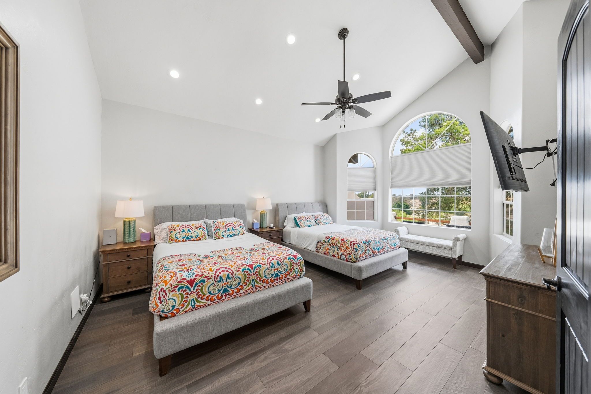 A bedroom with two beds, a ceiling fan, a TV mounted on the wall, and a large window with a view.