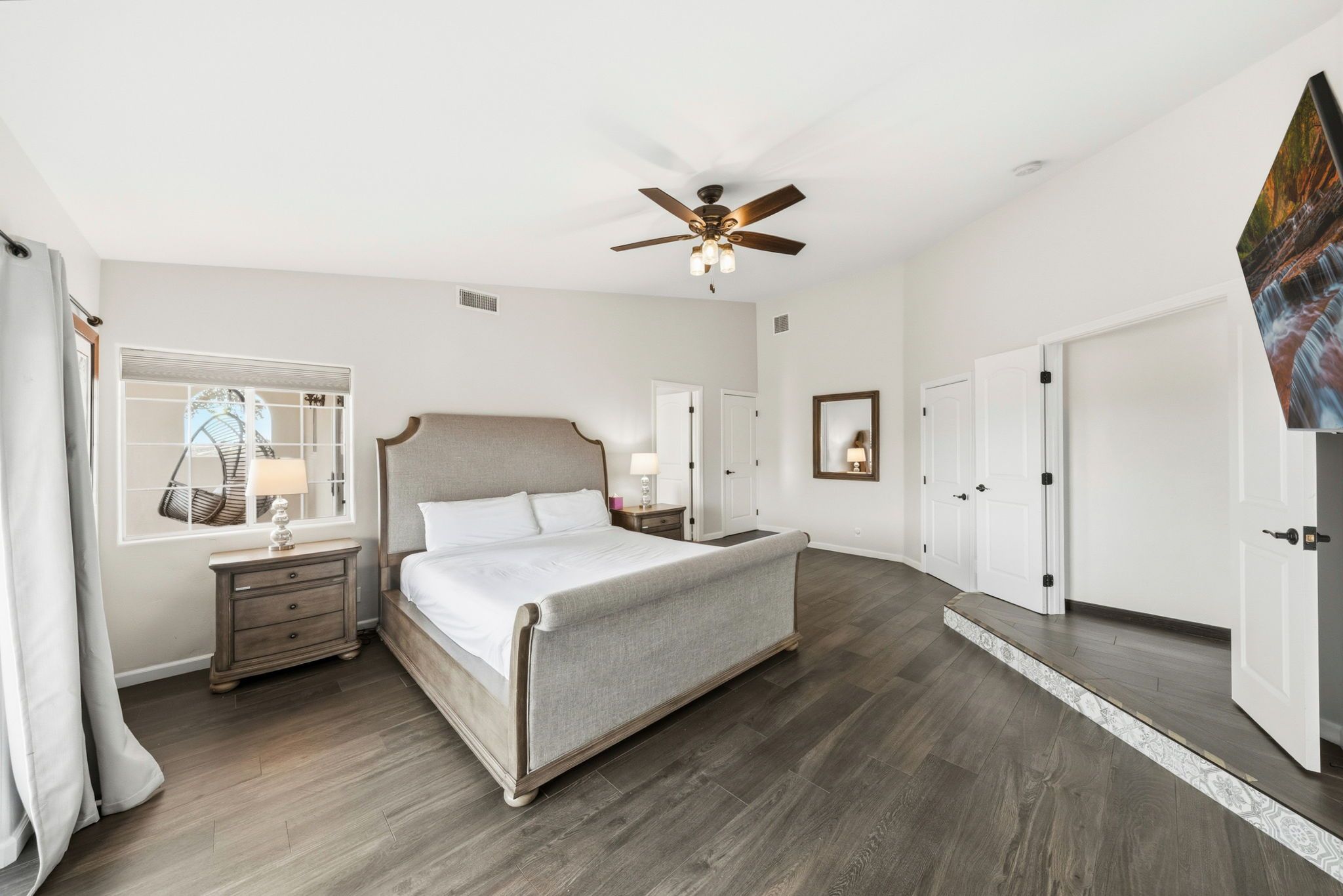 A bedroom with a large bed, a ceiling fan, a window with a view, and a door.