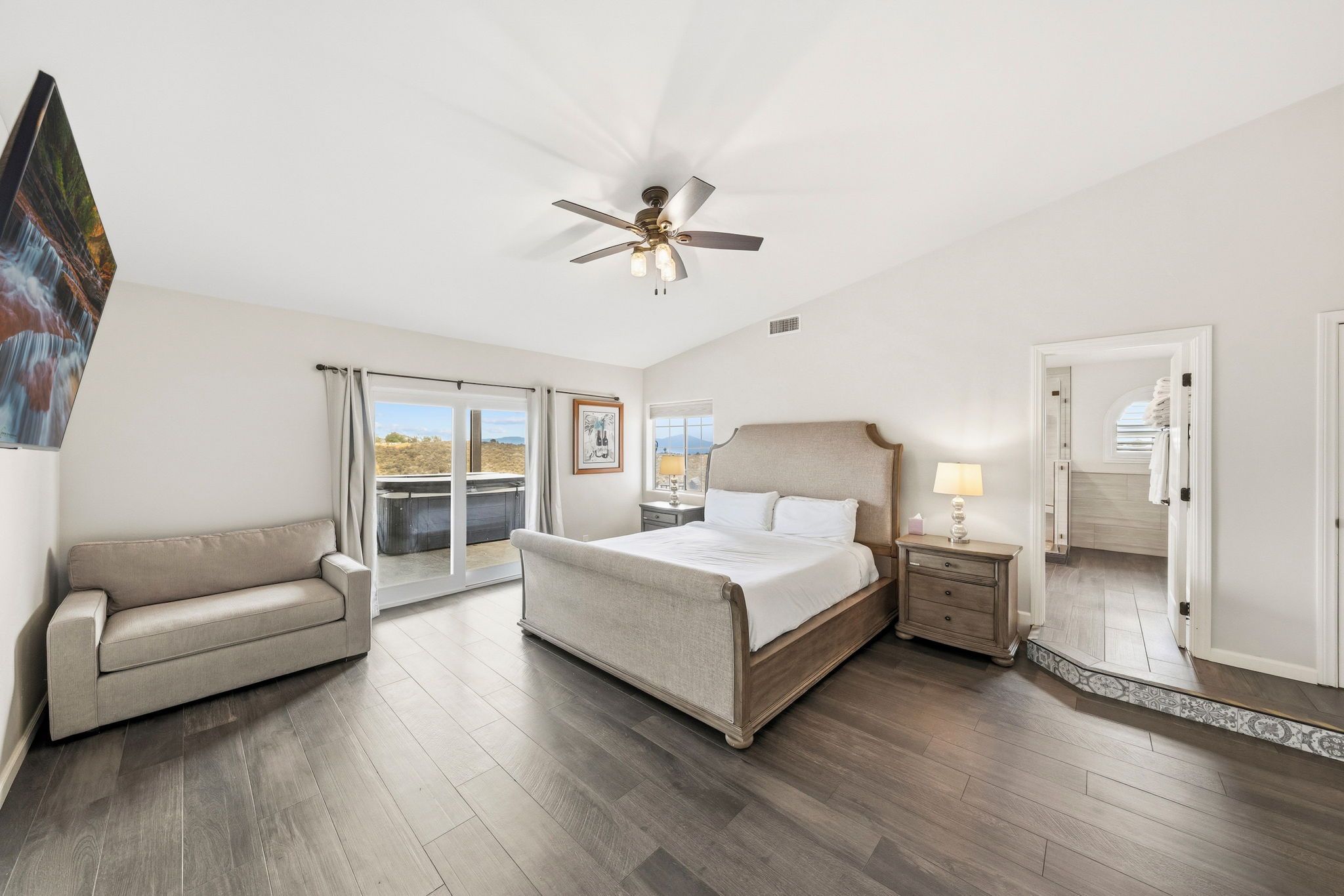 A bedroom with a bed, a sofa, a nightstand, a ceiling fan, and a view through a window.
