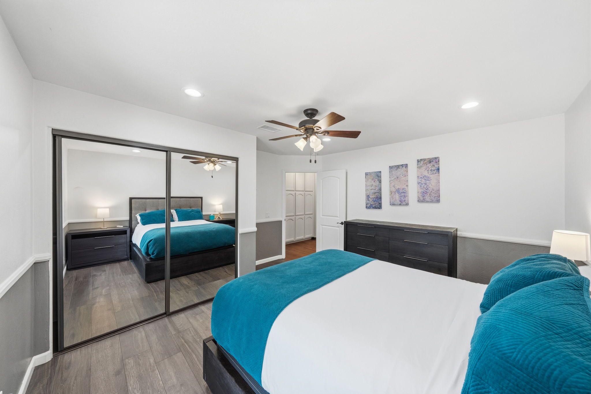 Une chambre à coucher comprenant un lit, une commode, un miroir, un ventilateur de plafond et des œuvres d’art accrochées au mur.