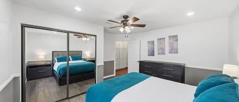 Une chambre à coucher comprenant un lit, une commode, un miroir, un ventilateur de plafond et des œuvres d’art accrochées au mur.