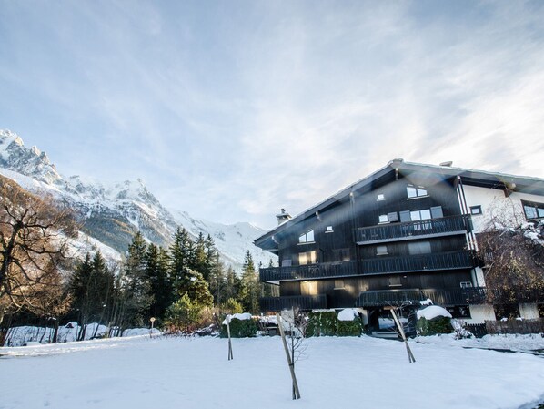Un edificio moderno de varios pisos con balcones, rodeado de montañas nevadas y árboles.