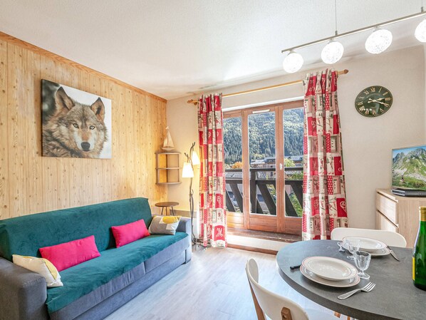 A cozy room with a green sofa, a painting of a wolf, a wooden wall, a dining table with plates and glasses, and a view of a mountain through a glass door. - Charming studio with Mont-Blanc view, Wifi and pets allowed (Chamonix-Mont-Blanc)