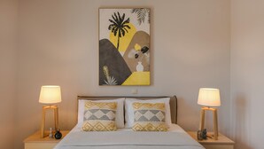 Un dormitorio con cama, dos mesitas de noche con lámparas, un cuadro colgado en la pared y una planta en la mesita de noche.