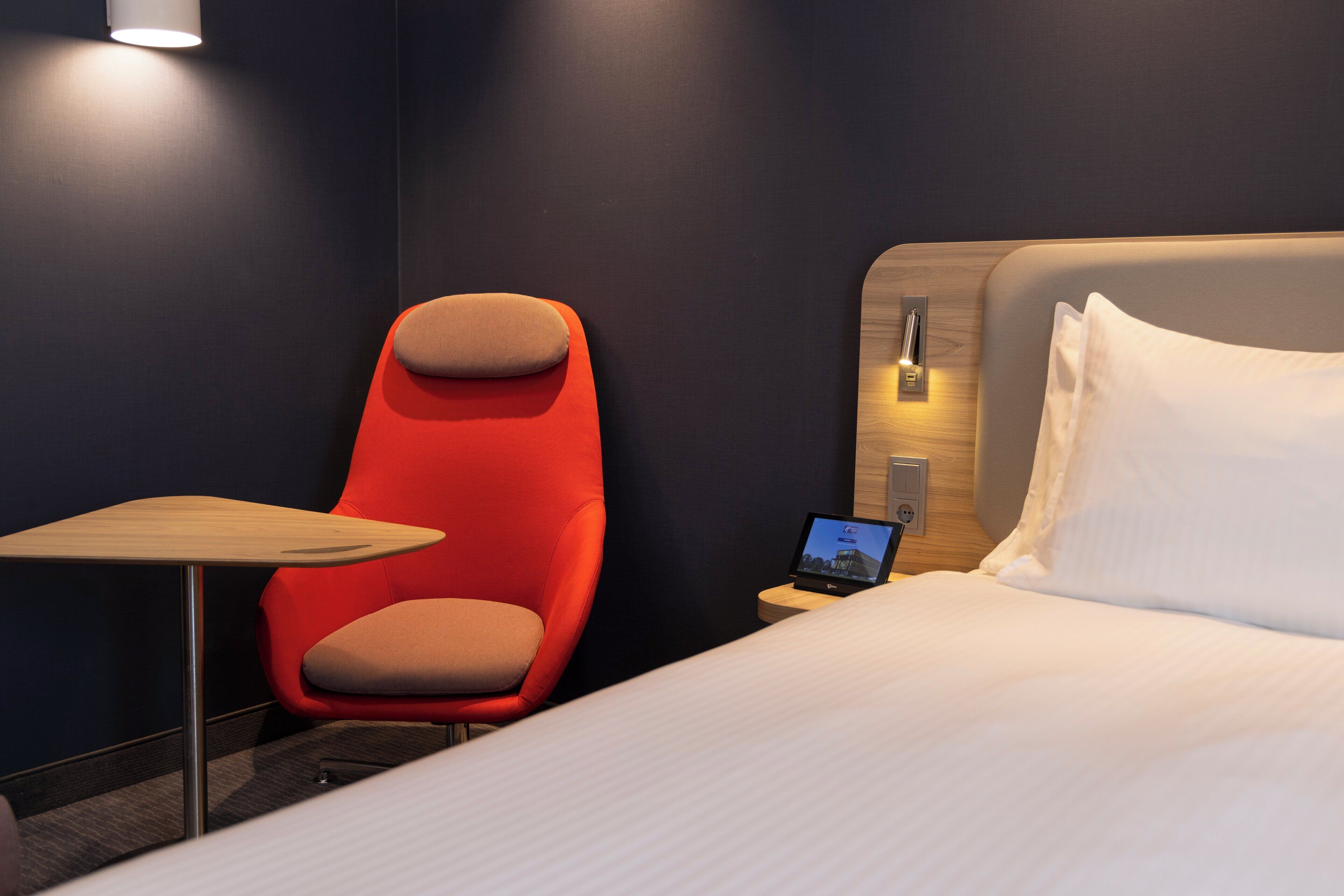 A hotel room with a bed, a red chair, a small table, and a laptop on a bedside table.