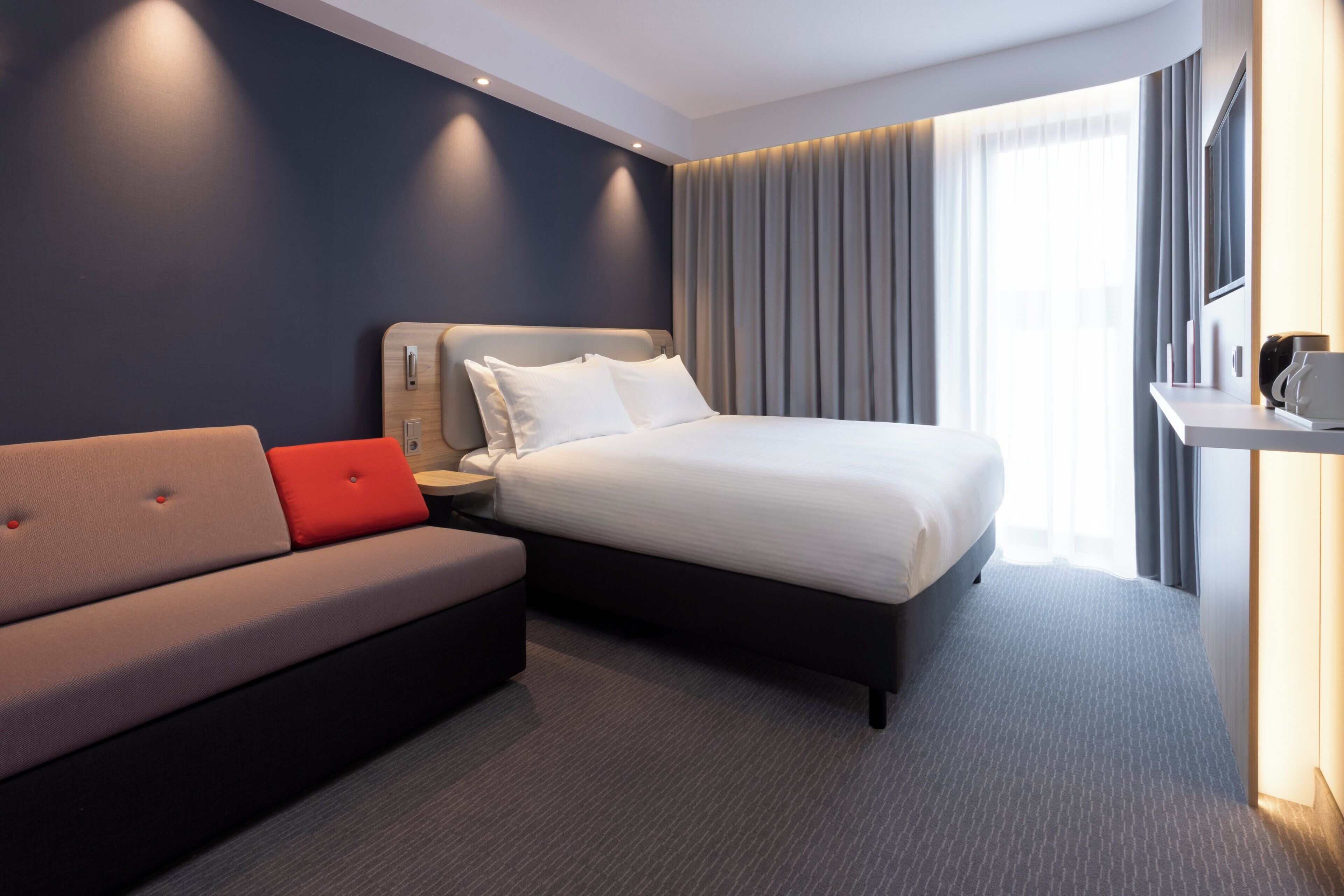 Una habitación de hotel moderna con cama, sofá, escritorio y un amplio ventanal con cortinas.