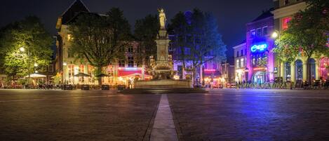 A nighttime city square with a central monument, cobblestone pavement, and surrounding buildings with illuminated signs.
