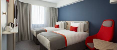 A hotel room with two beds, a red chair, a small table, a TV, and a window with curtains.