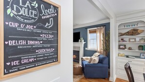 Un cartel de pizarra que enumera los favoritos locales y opciones gastronómicas en una sala de estar acogedora con un sofá azul, chimenea y estanterías blancas.