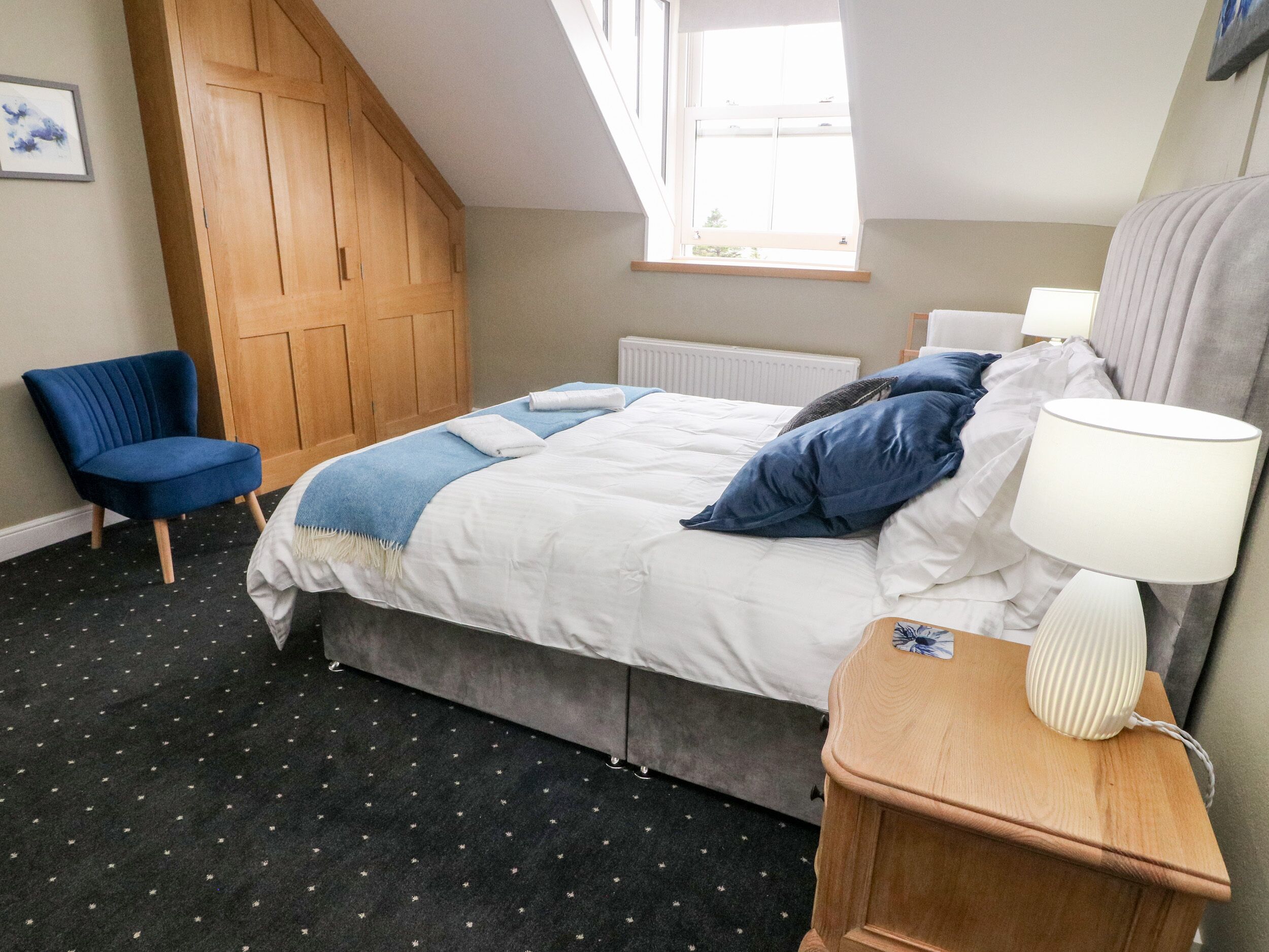 A bedroom with a bed, a chair, a bedside table with a lamp, a window, and a radiator.