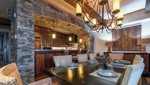 A spacious dining area with a large wooden table set for six, surrounded by cushioned chairs. A stone accent wall and a rustic ceiling add to the room's ambiance.